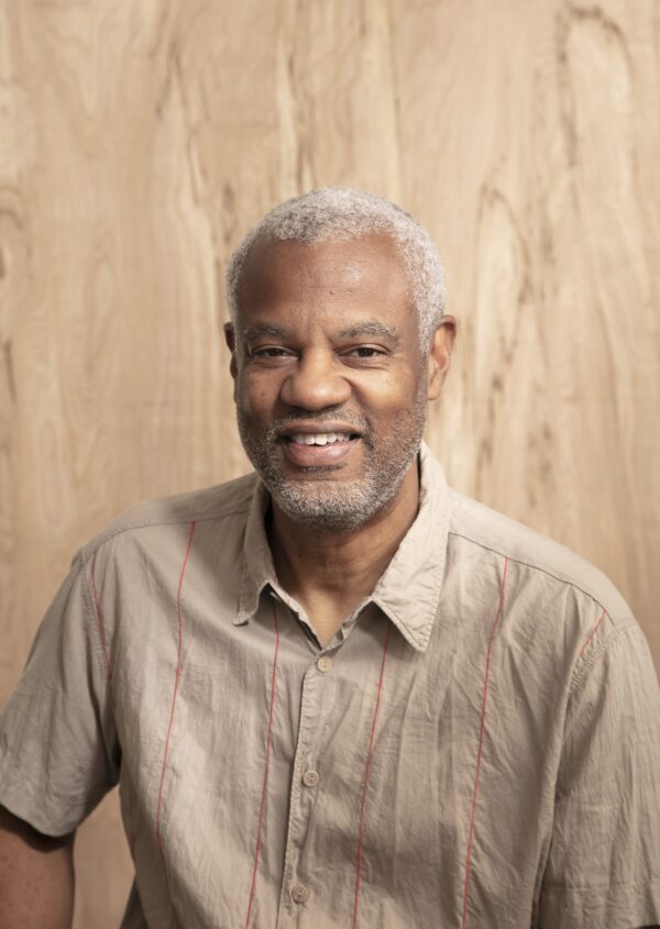 Portrait of a light brown skinned male with gray hair posed against a piece of wood. Their shirt matches the wood tone.