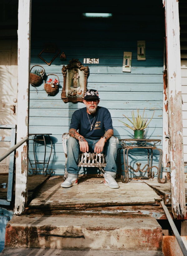 A person sits on a porch with weathered blue siding. The porch has decorative items on the wall, including a small statue in a frame. A plant in a pot is on a metal table nearby. The person wears casual clothes and a cap featuring the New York Yankees logo.