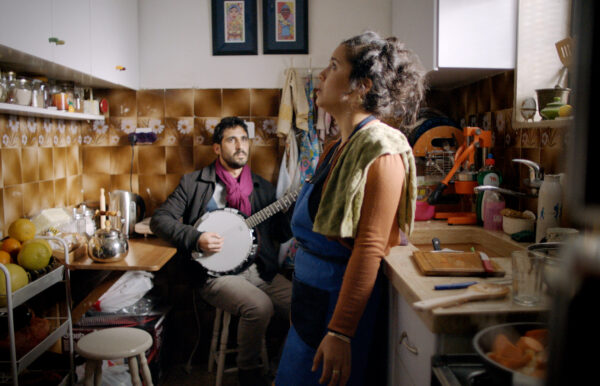 Two people in the middle of a small, well-used kitchen. One stands looking up at the ceiling while another holding a mandolin looks at the standing person.