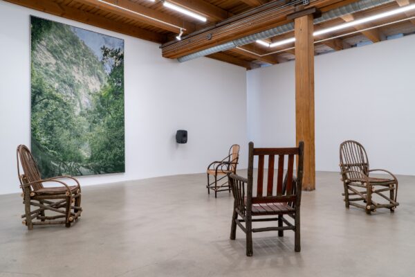 Installation in a gallery space featuring four wooden chairs in different styles placed in alternating directions and a single artwork of a green space hanging on a white wall