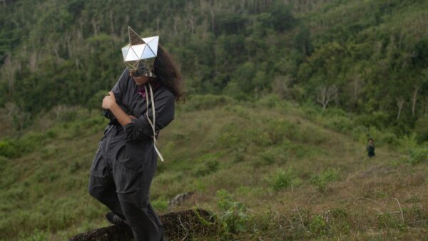 A person wearing a geometric, shiny silver mask that covers their eyes in the middle of a green field