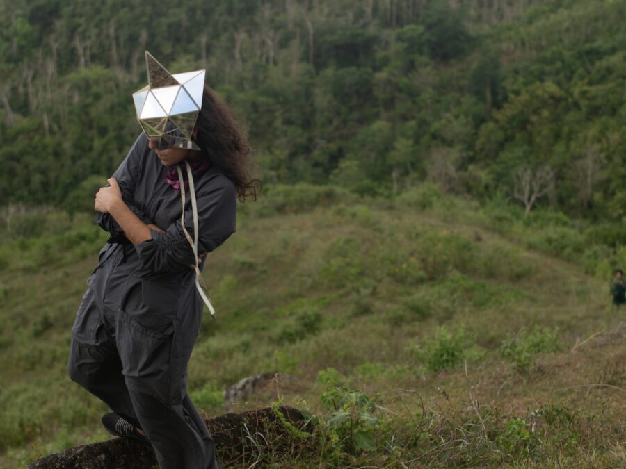 A person wearing a geometric, shiny silver mask that covers their eyes in the middle of a green field