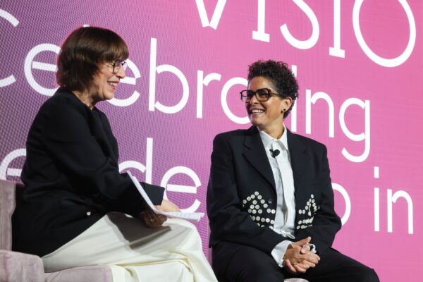 Two people sit smiling at each other in front of a large purple to pink screen displaying white text that is mostly blocked by their bodies