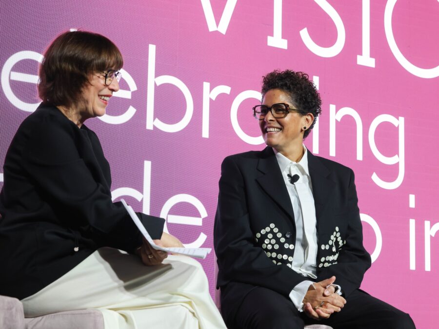 Two people sit smiling at each other in front of a large purple to pink screen displaying white text that is mostly blocked by their bodies