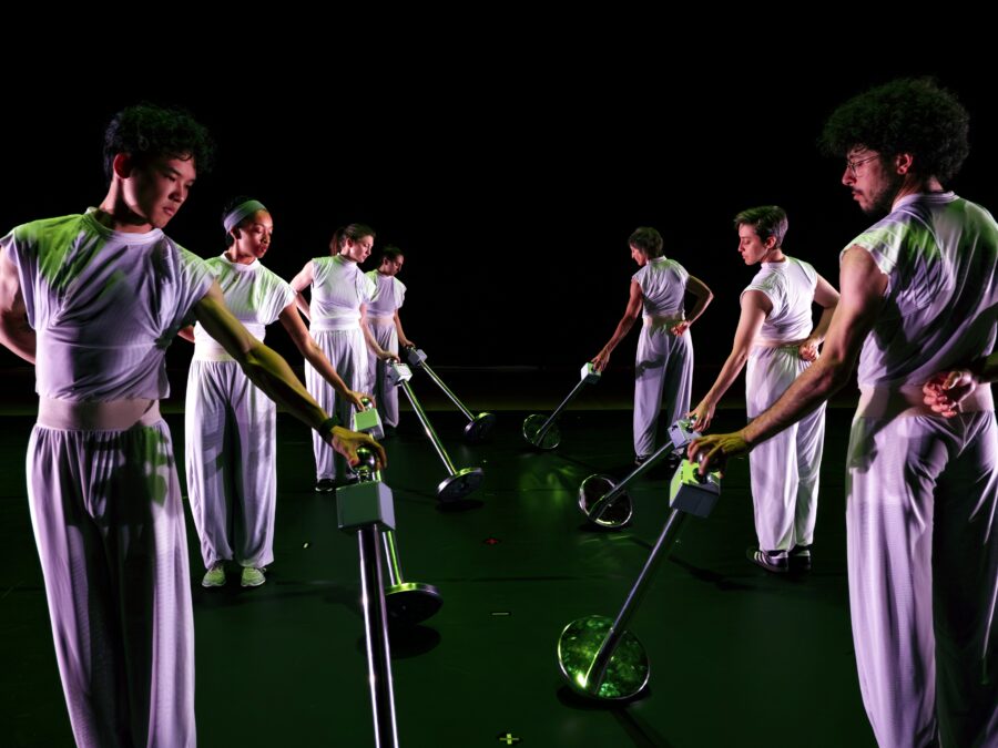 Two rows of people dressed in all white tilt stantions toward themselves on a dark stage