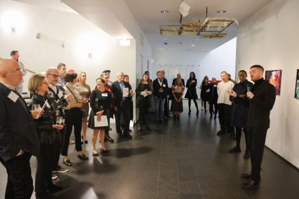 Large group of adults gathers around a wall with multiple framed artworks.