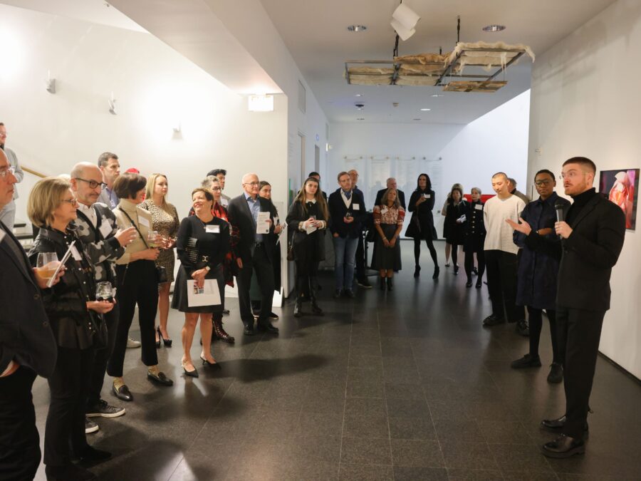 Large group of adults gathers around a wall with multiple framed artworks.