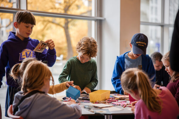 A group of kids sit around a table working on crafts in a room with floor to ceiling windows that feature trees with fall foliage