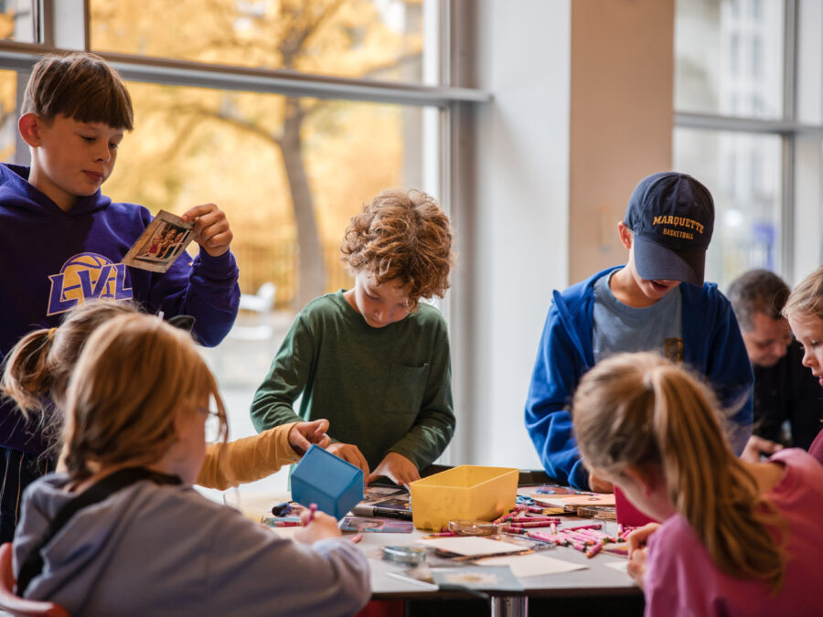 A group of kids sit around a table working on crafts in a room with floor to ceiling windows that feature trees with fall foliage
