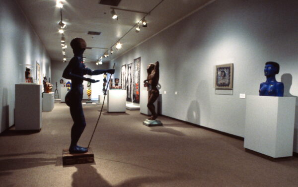 An art gallery displaying various statues and artworks. The gallery includes vibrant sculptures such as a prominent blue figure holding an object, and other abstract pieces are showcased throughout the well-lit space.