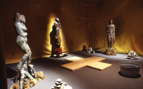 This image shows an art installation that features several human-like statues with textured surfaces, arranged around a cross-shaped wooden platform on the floor. The walls are lined with dark yellow fabric, adding to the dimly lit, contemplative atmosphere.