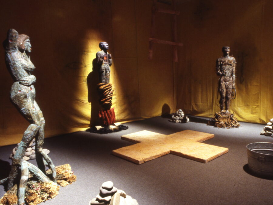 This image shows an art installation that features several human-like statues with textured surfaces, arranged around a cross-shaped wooden platform on the floor. The walls are lined with dark yellow fabric, adding to the dimly lit, contemplative atmosphere.
