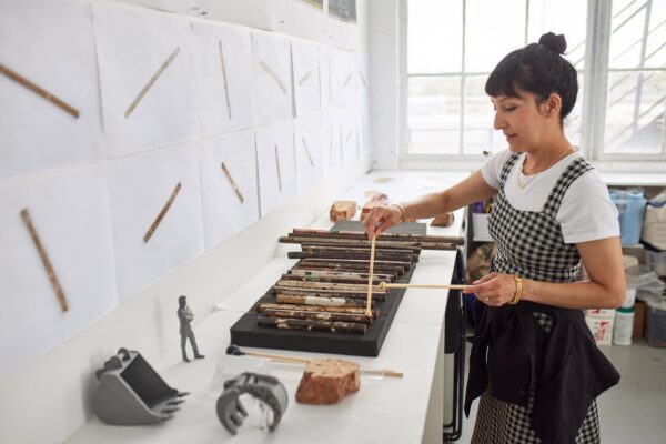 A female-presenting person plays a xylophone made out of rusty metal bars in a bright white room filled with scattered objects and images of the individual metal pieces printed on white paper