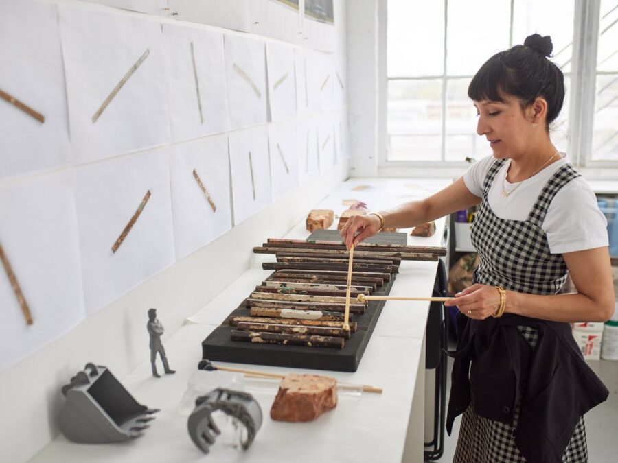 A female-presenting person plays a xylophone made out of rusty metal bars in a bright white room filled with scattered objects and images of the individual metal pieces printed on white paper