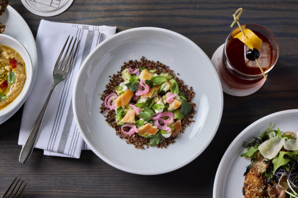A grain salad in a white bowl on a wooden table next to a napkin and fork, an old-fashioned cocktail, and two other dishes in white dishes