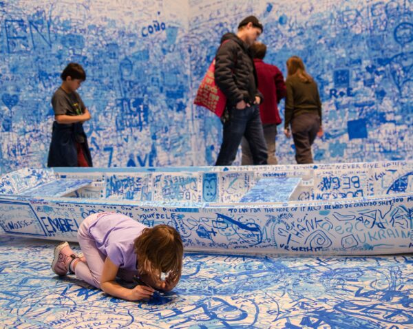 A child writes on the floor in a room filled with blue writing all over the white walls and boat. Other people mill about in the background.