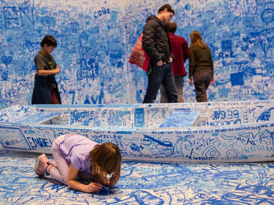 A child writes on the floor in a room filled with blue writing all over the white walls and boat. Other people mill about in the background.