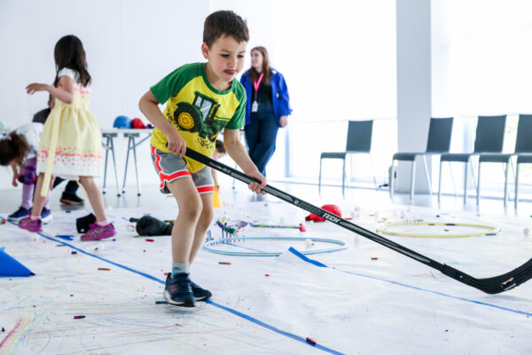 A child plays indoor hockey in a bright white room with other children and adults nearby