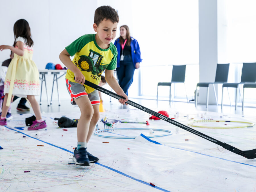 A child plays indoor hockey in a bright white room with other children and adults nearby