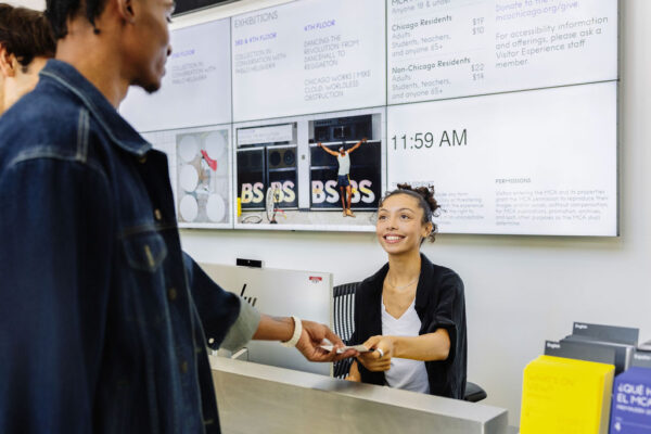 A young woman behind a computer and sitting in a desk chair smiles as she hands two young men tickets.