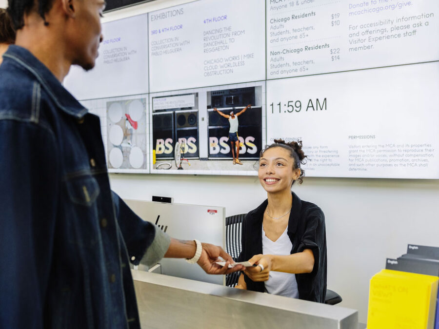 A young woman behind a computer and sitting in a desk chair smiles as she hands two young men tickets.