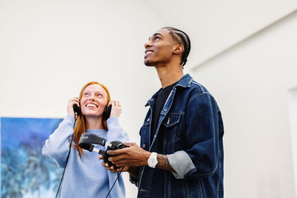 Two young adults smile as they look up at something not pictured. There is a large artwork behind them with shades of blue.