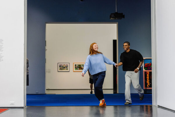 Two young adults hold hands as they run through a gallery space.