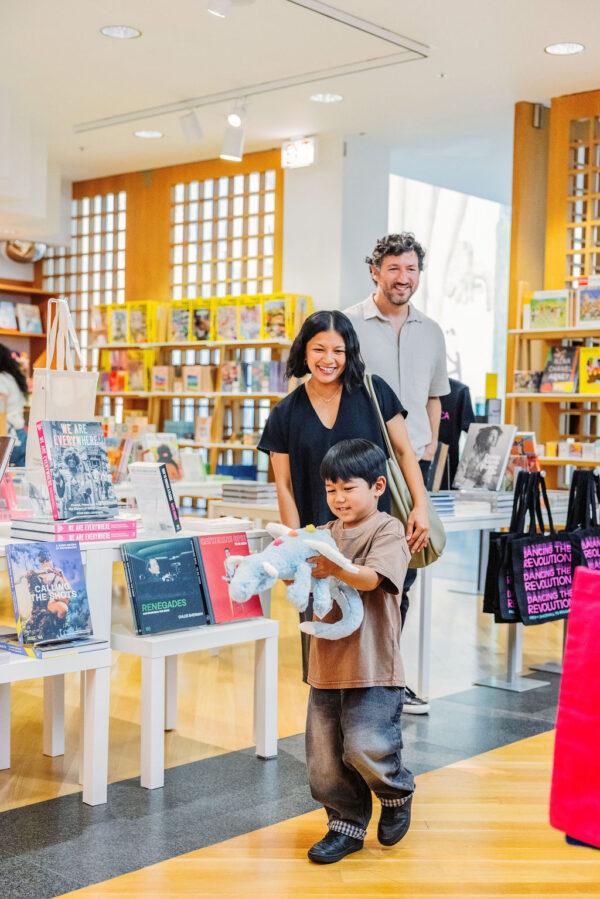 A young boy walks in front of a woman and man, smiling, as he holds a stuffed animal.