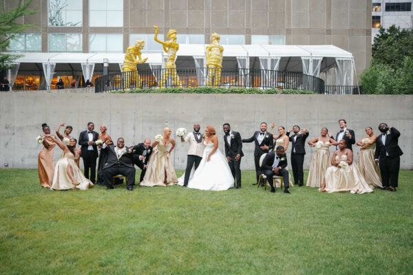 A wedding group poses in a garden below three yellow figures near a rental tent