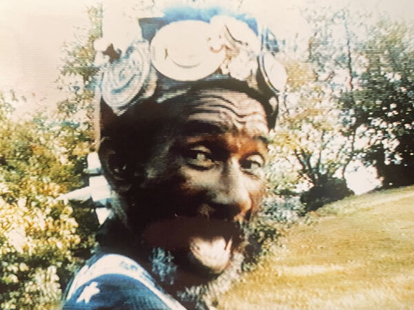 A grainy image of a Black man turning toward the viewer with their mouth open in surprise or excitement. They wear a hat covered in circular metal objects.