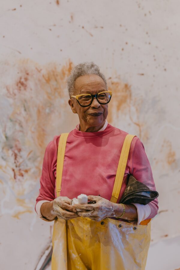 A person in yellow waders and a salmon-colored shirt holds a large piece of chalk that has rubbed off on their hands. Behind them abstract brown splashes of paint cover the middle portion of a white wall.