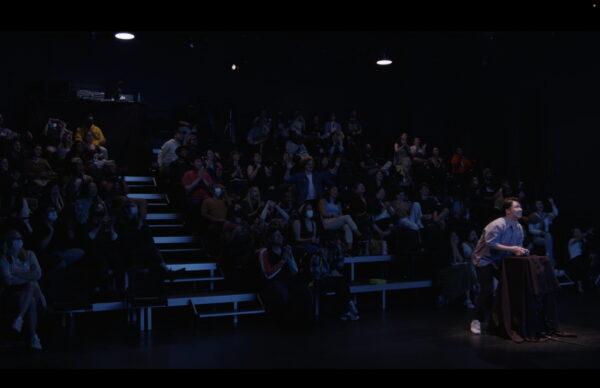 An audience cheers with their hands in the air, supporting a player with a PlayStation controller on stage. The player is gritting their teeth and smiling in tense focus.