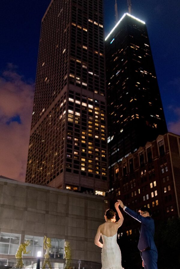 A woman in a white wedding dress and a man in a navy suit dance in the MCA’s backyard with tall Chciago skyscrapers including the John Hancock building as the backdrop
