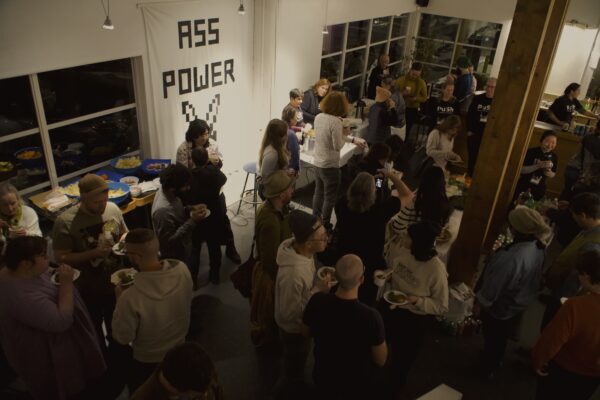 A lobby in a theatre filled with people holding small cups of soup and plates of falafel. A banner hangs on the wall that says “Ass Power” and there are large bowls of chips and snacks on a nearby table.
