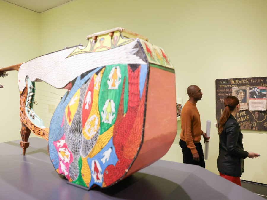 A large, colorful boat-like instrument takes up a large portion of a key lime colored room, behind which two people look at an artwork on the wall filled with writing, including Lee Scratch Perry’s name.