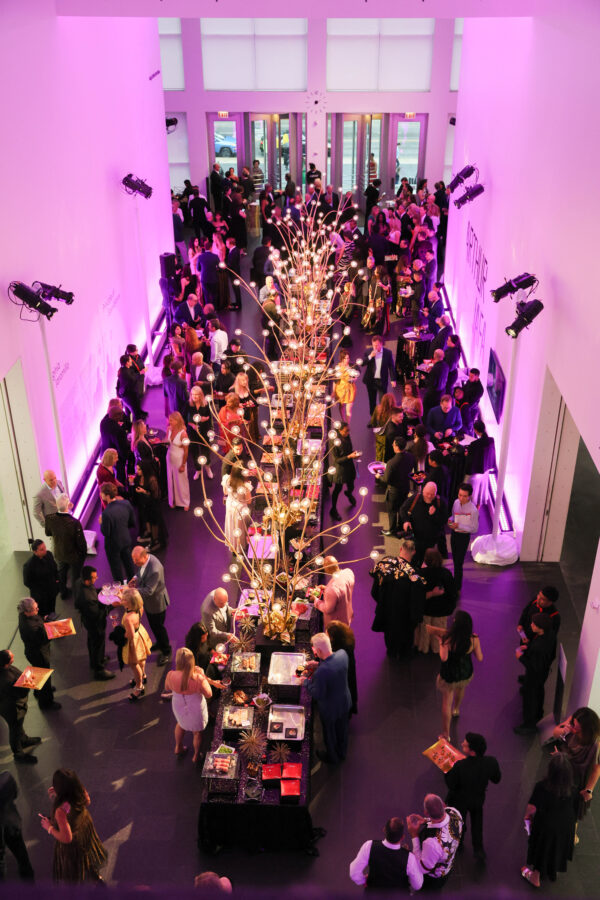 Overhead shot of a crowd mingling around a long buffet table in a two story event space lit with purple lights