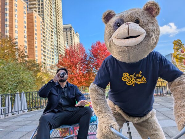 A taupe bear mascot in a blue Bad at Sports t-shirt leans into the camera as a person in all black leans back, their face hidden by a glass bottle that they are taking a sip out of