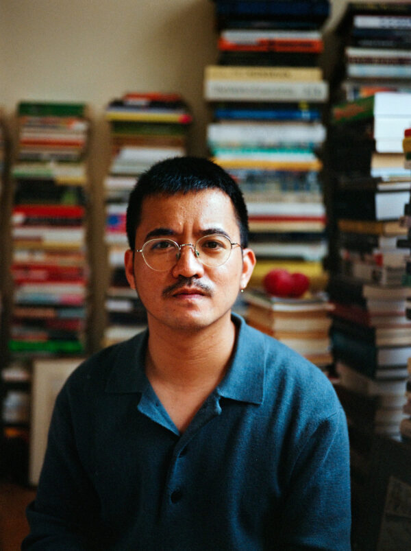 Photo of man wearing glasses and blue shirt looking at the camera and sitting in front of several stacks of books.