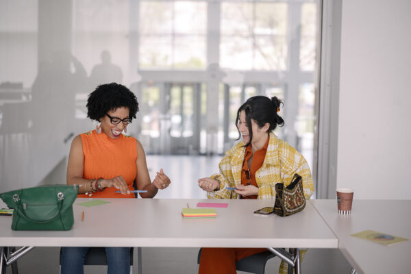 Two people smile while working on a project together in a bright white space with a window behind them looking down onto a bright atrium