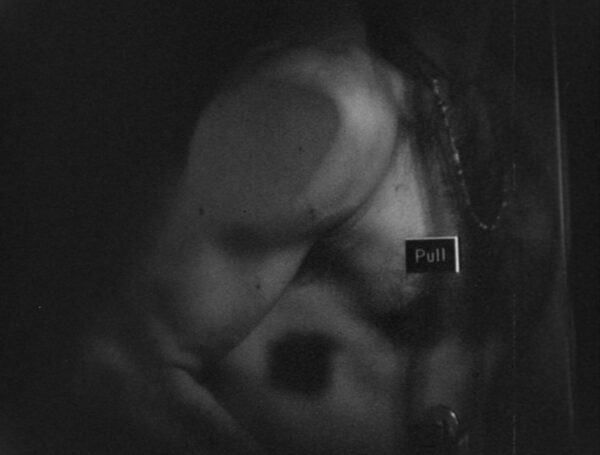 A dark black and white image of a nude male torso pressed up against a piece of glass with a small placard labeled pull, suggesting a door