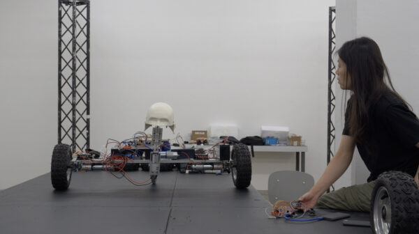A person kneels on the floor, connecting wires to circuit boards, next to a robotic vehicle. The vehicle, on a black platform, has four large wheels, exposed electronics, and a white skull mounted on top.