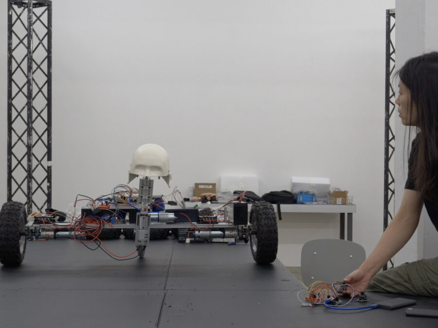 A person kneels on the floor, connecting wires to circuit boards, next to a robotic vehicle. The vehicle, on a black platform, has four large wheels, exposed electronics, and a white skull mounted on top.