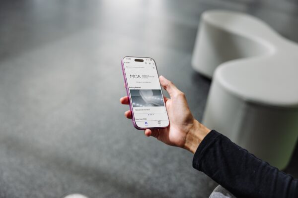 A right hand is extended over a grey floor while holding a phone displaying an image of the MCA staircase. A grey bench in the form of a splat appears to the right.