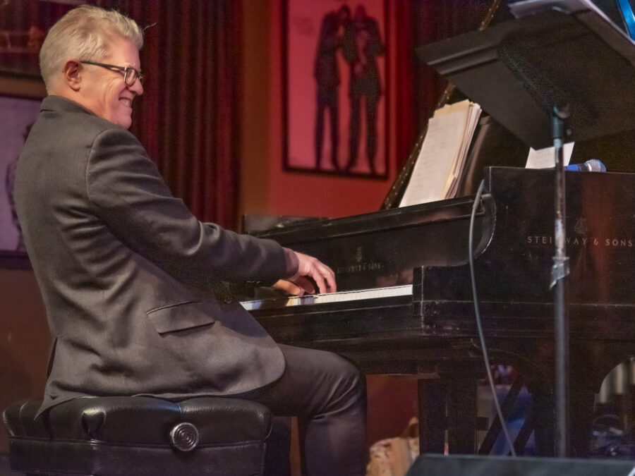 A male-presenting person in a grey suit plays a black grand piano on a red-lit stage. His face expresses he is into the groove of the music.