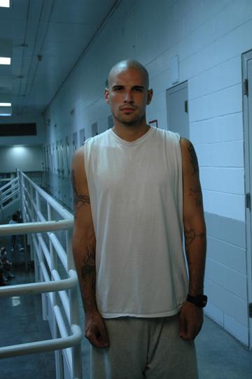 Tattooed person with mustache dressed in white tank top and grey sweatpants stands on the mezzanine of a prison looking at camera.