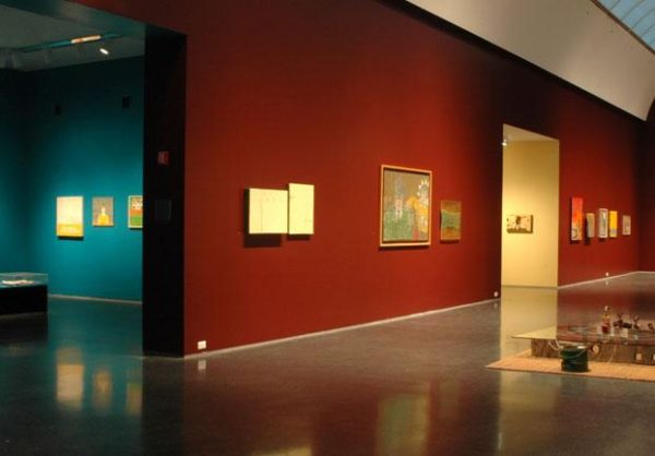 An installation view shows artworks hanging on the red-painted walls of a gallery, with another turquoise gallery to the side.