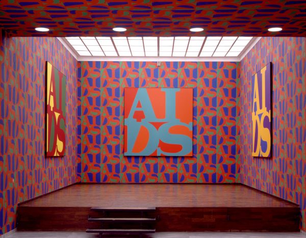 An elevated room covered in red, blue, and green repeating wallpaper on three walls that matches the art which features the acronym AIDS in the style of the Robert Indiana LOVE artwork on the three walls.