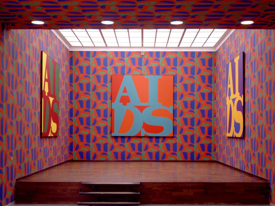 An elevated room covered in red, blue, and green repeating wallpaper on three walls that matches the art which features the acronym AIDS in the style of the Robert Indiana LOVE artwork on the three walls.