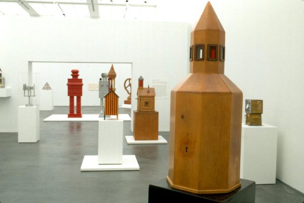 Several wooden sculptures on pedestals in a white room. Some resemble rocket ships, others resemble human figures.
