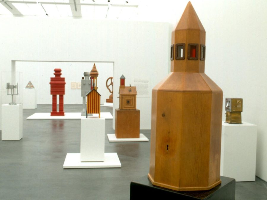 Several wooden sculptures on pedestals in a white room. Some resemble rocket ships, others resemble human figures.